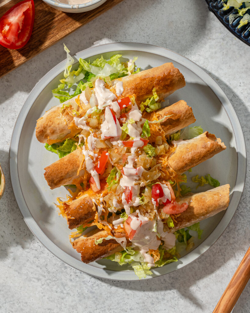 A plate of low carb taquitos with sauce and toppings over a gray marble surface.